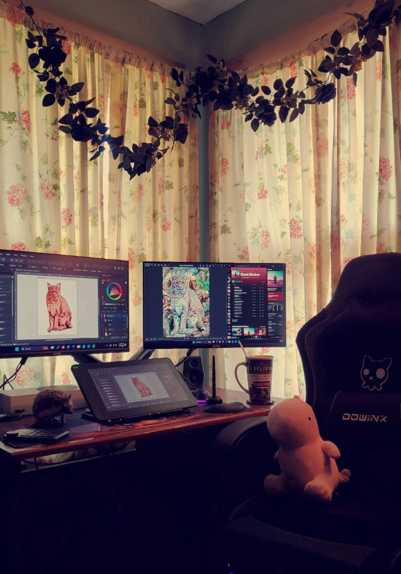 Creative design setup featuring a dual-monitor workspace with a drawing tablet, Spotify and digital art software open, and a hedgehog posing on the desk beside a plush toy in the chair.