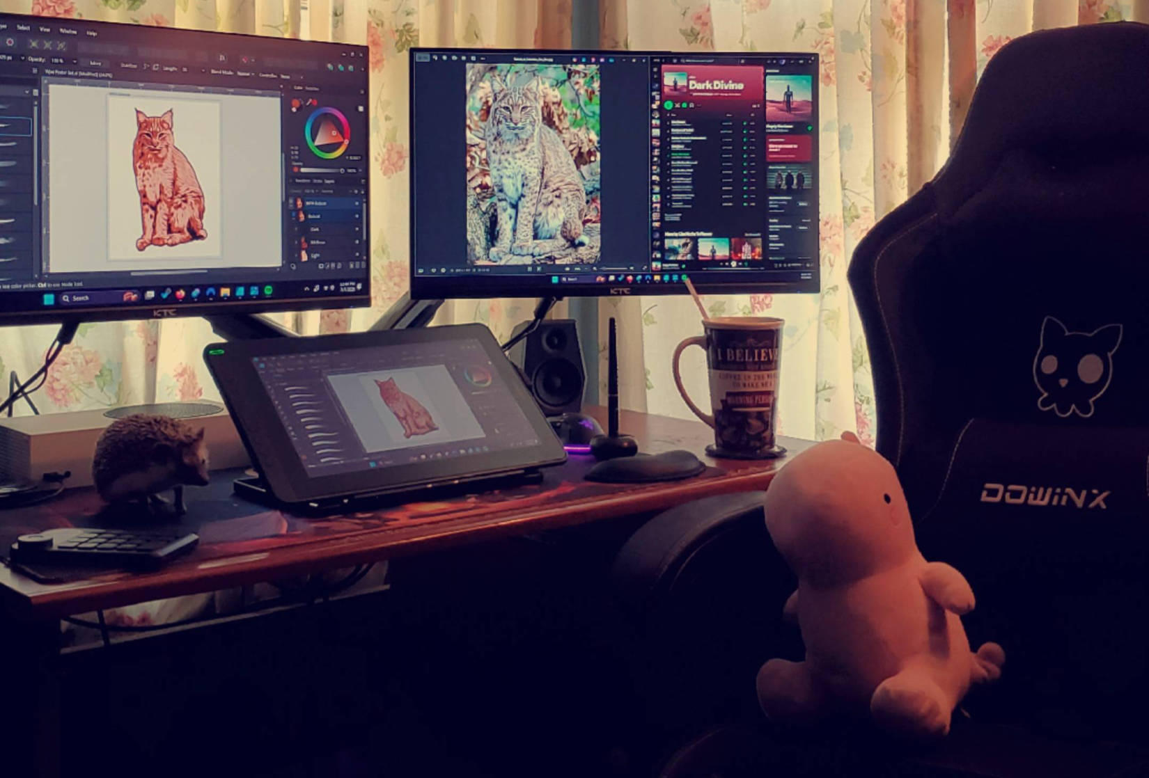 Creative design setup featuring a dual-monitor workspace with a drawing tablet, Spotify and digital art software open, and a hedgehog posing on the desk beside a plush toy in the chair.