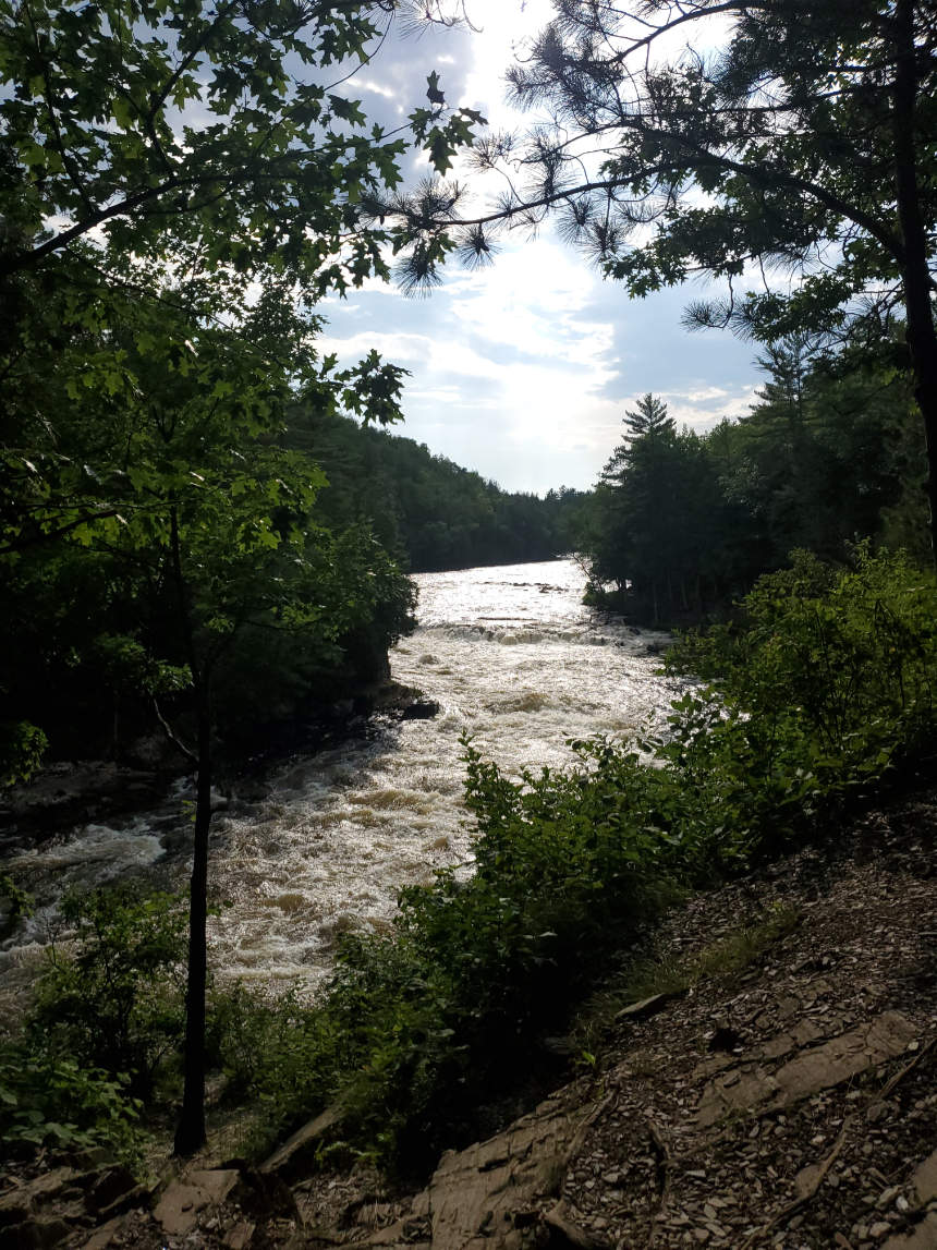 A midday view of the winding Menominee River flanked by bright green foliage in a Wisconsin landscape. The calm river snakes through the vibrant forest, captured just before a white water rafting adventure.