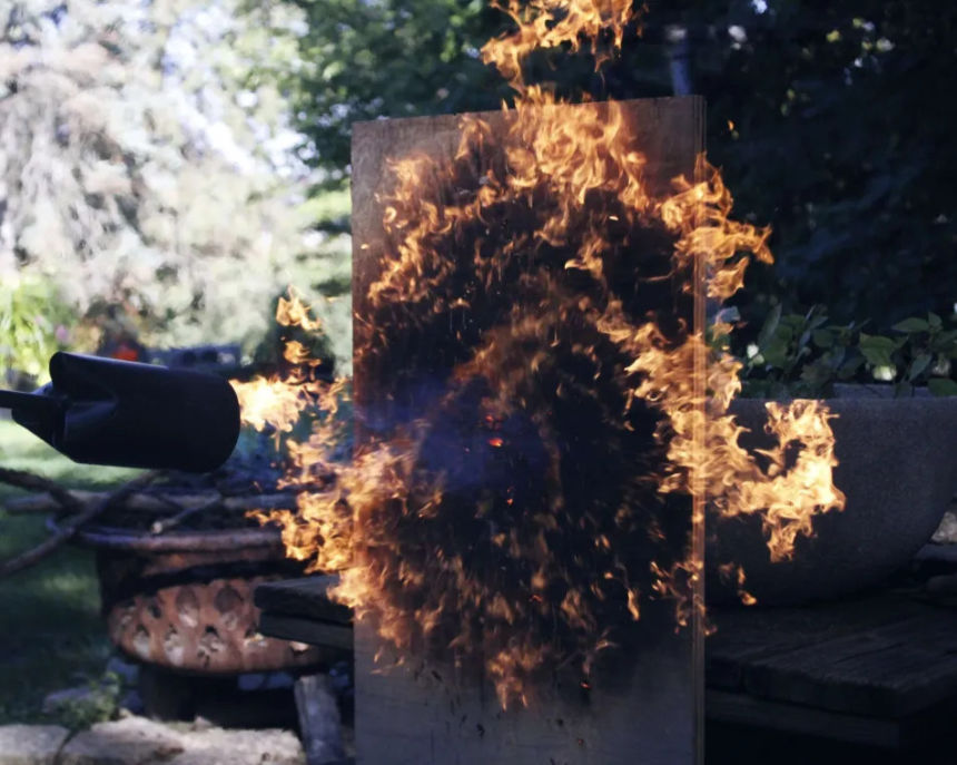A propane flamethrower shoots flames shaped like a dancing maple leaf onto a wooden board, showcasing dynamic fire movement and intricate details. The fiery display contrasts sharply with the lush green trees in the background.