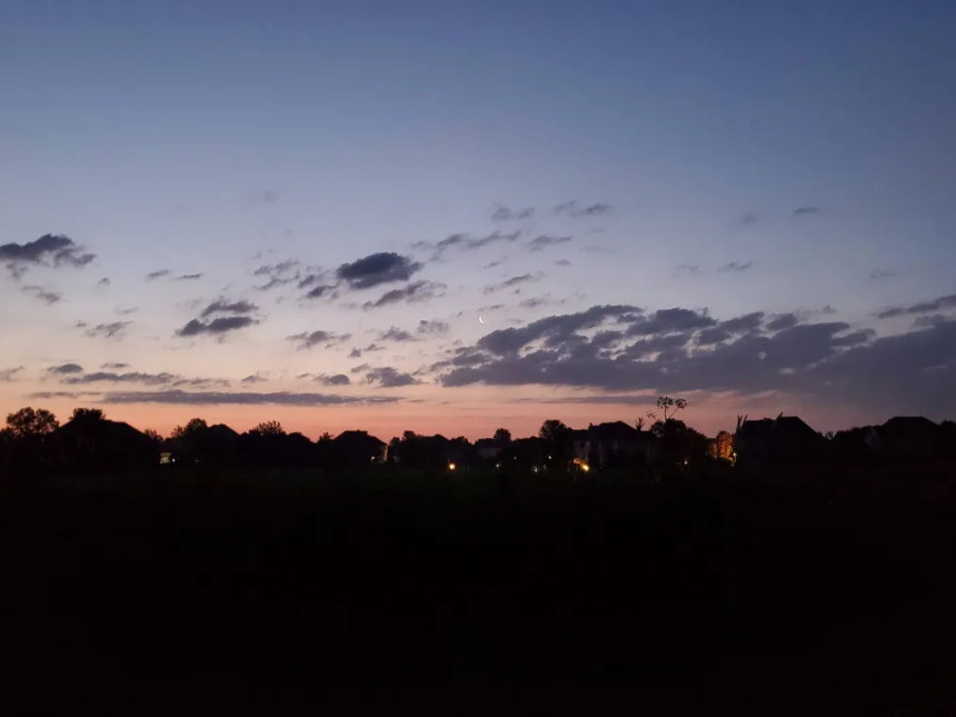 Silhouetted houses stand quiet in the early morning light, their dark outlines broken only by the soft glow of a few street lamps and faintly lit windows. Above them, a brilliant sunrise paints the sky in a gradient of orange-pink, fading into pale blue and then deep navy—capturing the stillness and beauty of dawn.